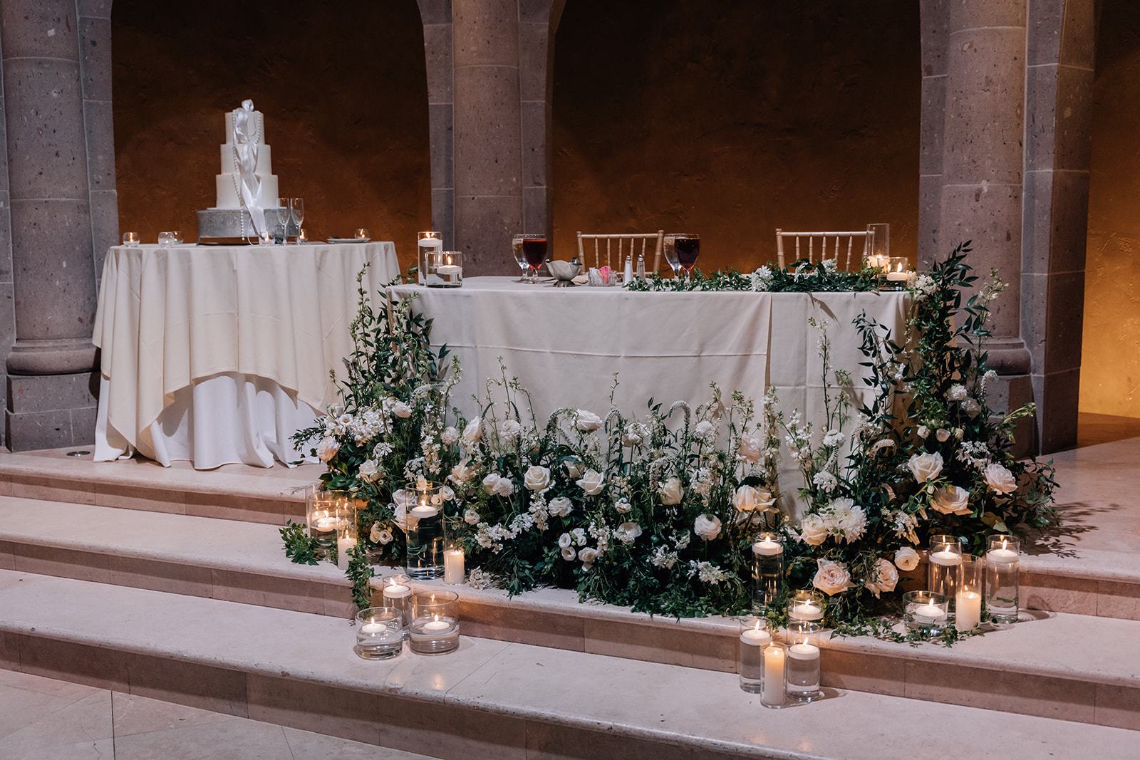 Candlelit sweetheart table trailing white garden roses and greenery in a stone hall