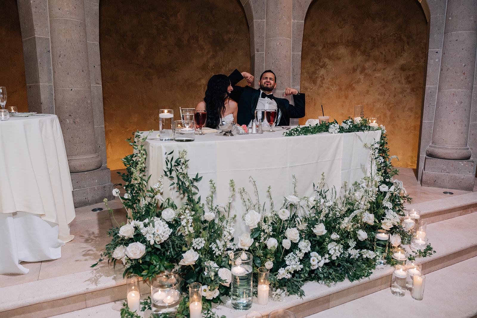 Newlyweds at a garland-draped sweetheart table in a stone reception hall